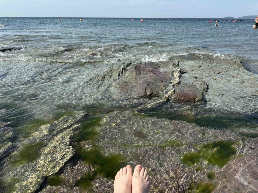 Füsse am Meer im Wasser. Im Hintergrund Felsen, Meer und blauer Himmel.